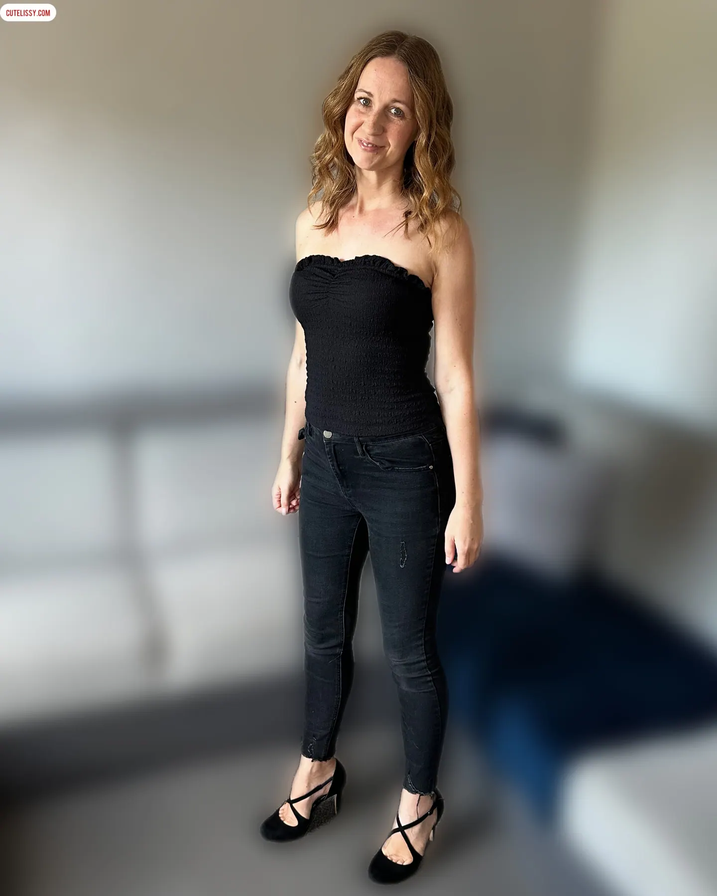 Lissy standing and smiling in a black strapless top, black jeans and heels.