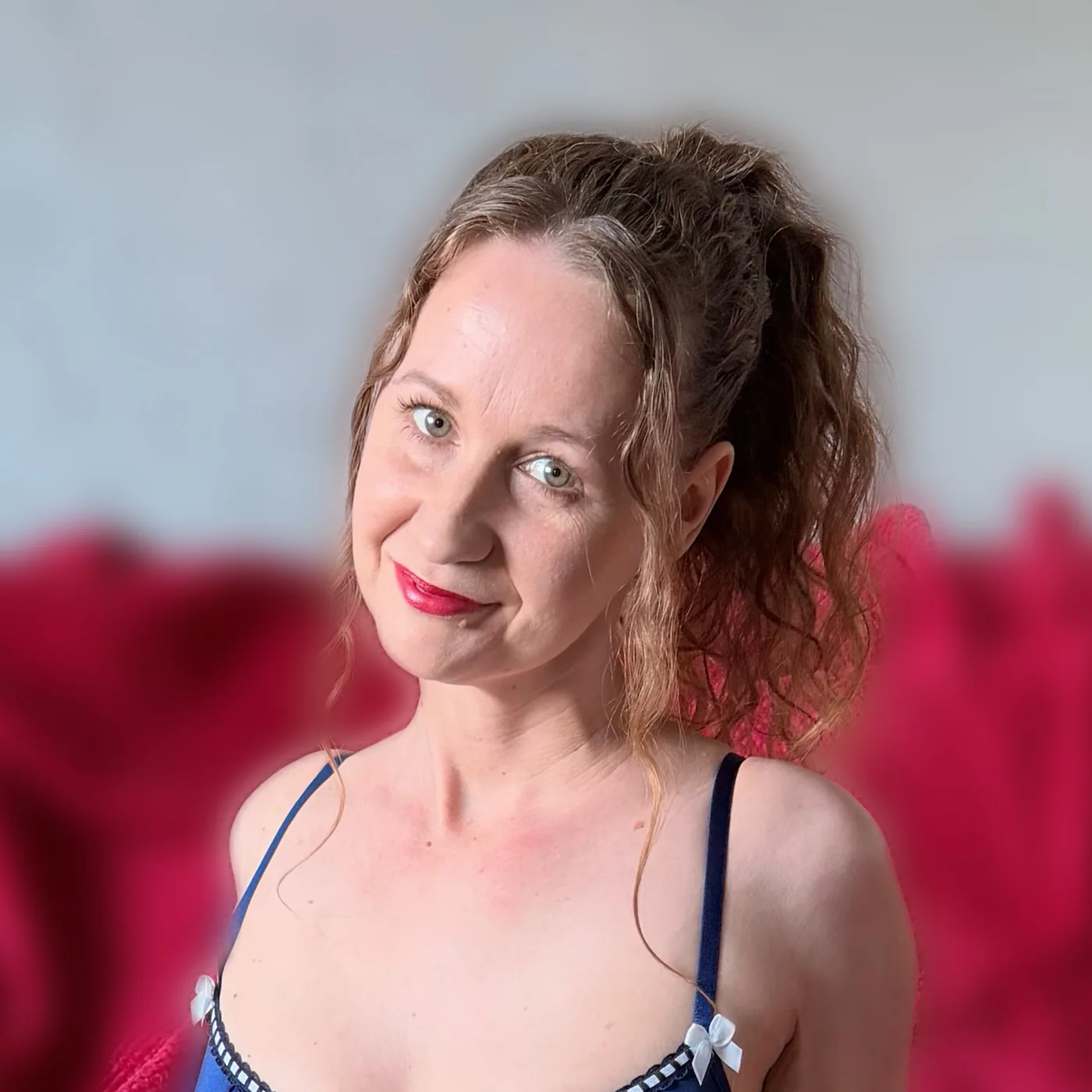 Lissy smiling over her shoulder in a navy blue lace bodysuit, with a deep red throw behind her.